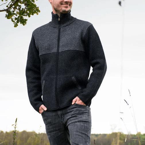 Ein Mann steht draußen auf einer Wiese. Er trägt eine zweifarbige Strickjacke in Grau und Schwarz mit Reißverschluss, hohem Kragen und Taschen. Dazu eine graue Jeans. Er schaut lächelnd nach links.